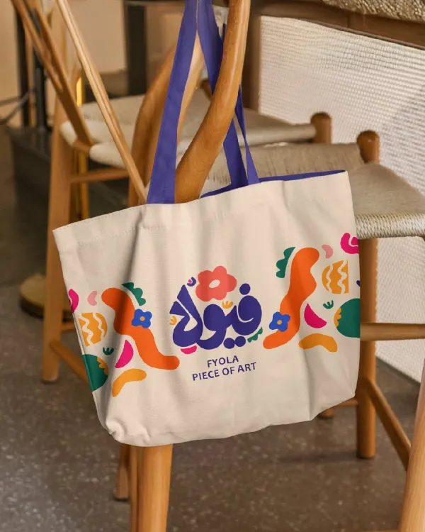 Create a professional lifestyle merchandise mockup for the uploaded brand assets. The scene features a high-quality canvas tote bag hanging casually from the back of a modern wooden chair in a stylish café or interior setting. The front of the tote bag prominently displays a vibrant and artistic collage design, automatically incorporating the uploaded brand's logo by abstract, organic shapes and colorful patterns that match the brand's visual identity. The handles of the bag feature a bold contrast color derived from the brand's palette. The background shows a warm, contemporary architectural space with additional minimalist chairs and tables softly blurred to maintain focus on the product. Warm, natural indoor lighting casts gentle shadows, emphasizing the rich texture of the canvas fabric and the smooth wood of the chair. The shot is captured from a slightly close-up, natural eye-level angle, creating an elegant, modern, and premium lifestyle aesthetic.