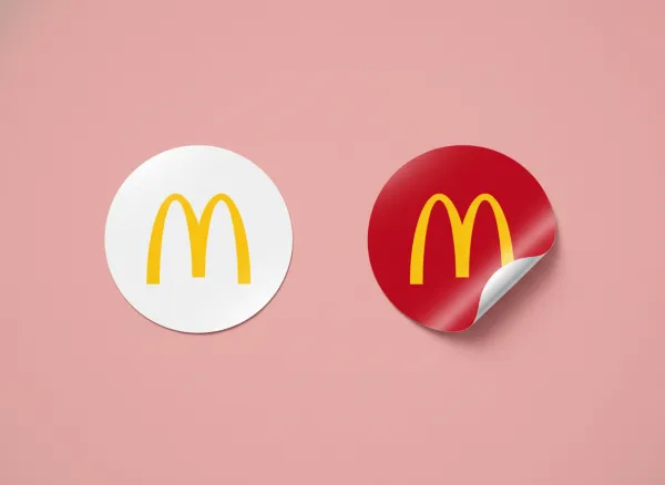 Photorealistic mockup on a simple background in a slightly lighter shade of the brand’s primary color. Show two circular stickers, both featuring the uploaded logo: one white sticker flat on the surface, and one brand-color sticker slightly peeling. Include realistic shadows, subtle reflections, and slight texture for a natural, tactile look. Clean, professional, and visually appealing.