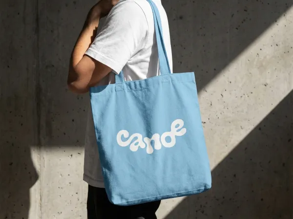 Apply logo to a brand-colored cotton tote bag carried by a person in a white T-shirt. Concrete wall background with sharp diagonal sunlight and shadows. Minimalist lifestyle photography, realistic fabric textures.