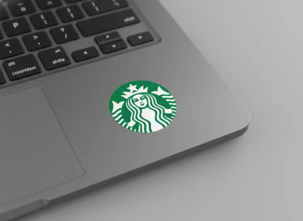 Extreme close-up of a laptop trackpad with a sticker prominently displayed. Sticker features the uploaded logo or brand text in brand colors. Laptop keys partially visible in the background. Bright, even lighting with minimal shadows. Top-down angled view emphasizing sticker details and placement. Clean, modern, photorealistic style on a plain gray background.