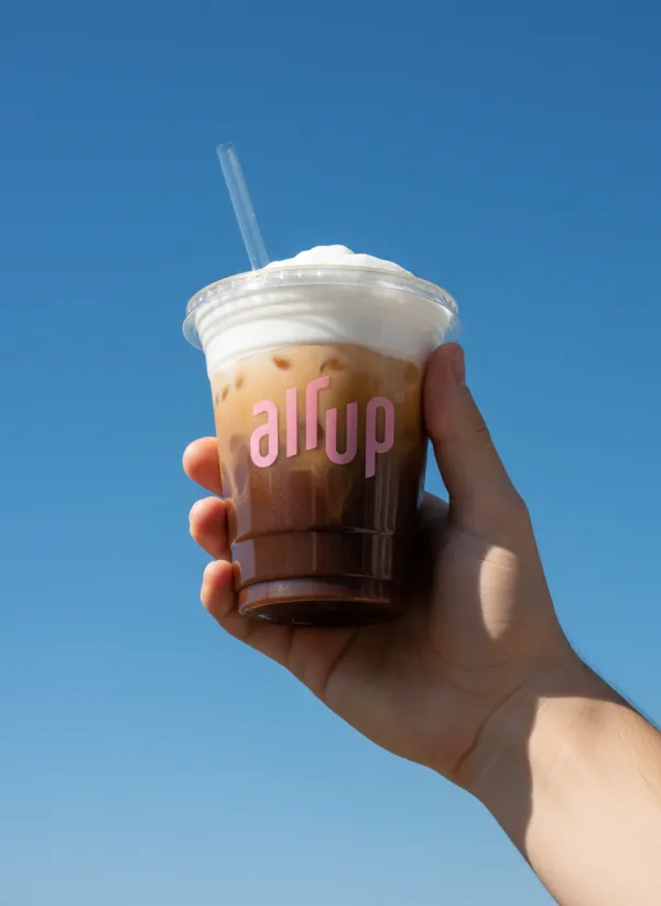 Apply the uploaded logo onto the side of a clear plastic takeaway coffee cup held aloft by a person's hand. The cup contains a layered iced coffee with a thick, creamy foam top and dark coffee base. The scene is set against a vibrant, solid clear blue sky background. Bright, direct sunlight creates a clean aesthetic with a soft shadow on the hand and crisp highlights on the plastic texture. Low-angle shot, professional lifestyle photography, minimalist composition, 8k, photorealistic.